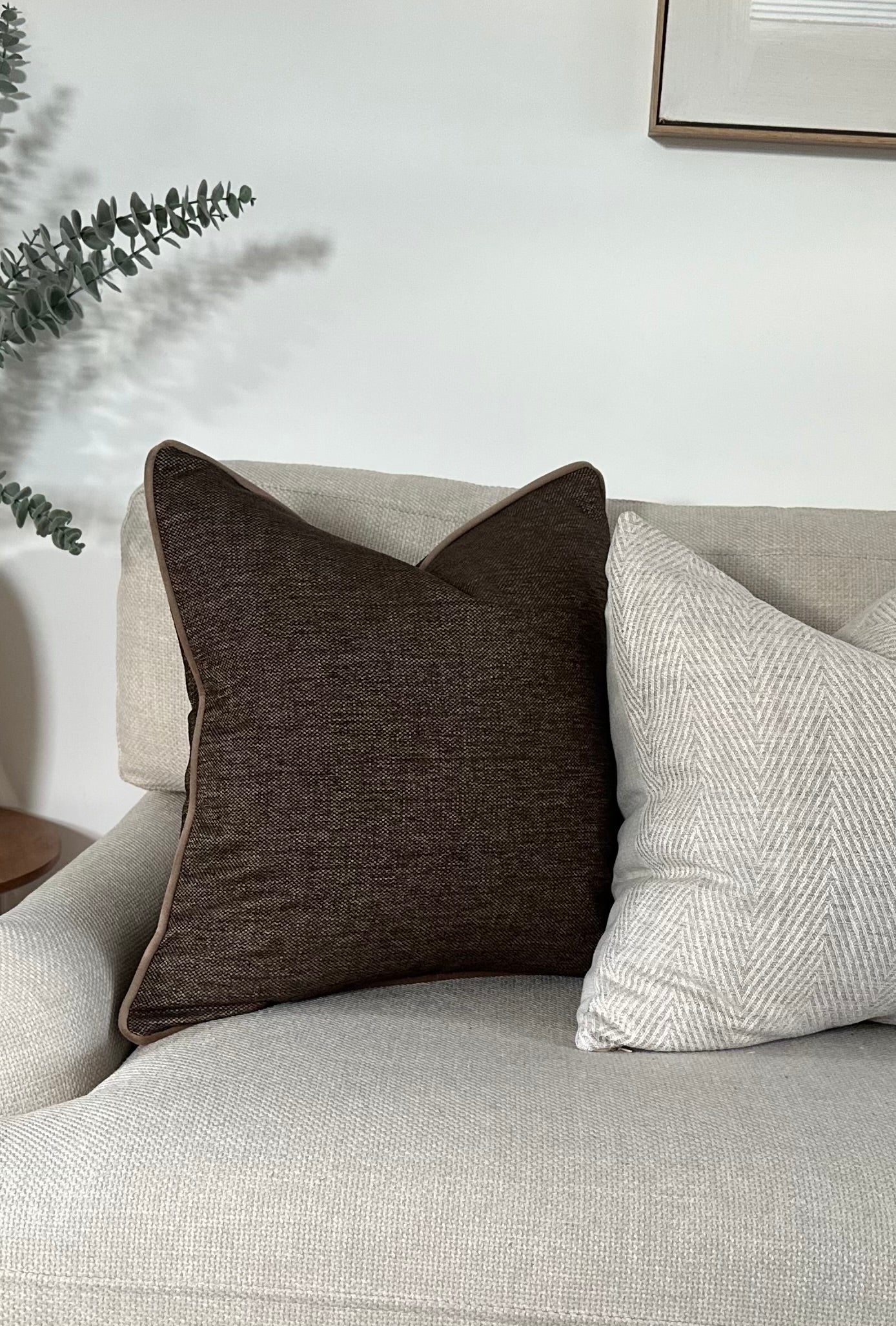 dark brown cushion cover with subtle textured fabric and mocha piping, styled on a neutral armchair beside a light herringbone pillow. Handmade, luxury feel.