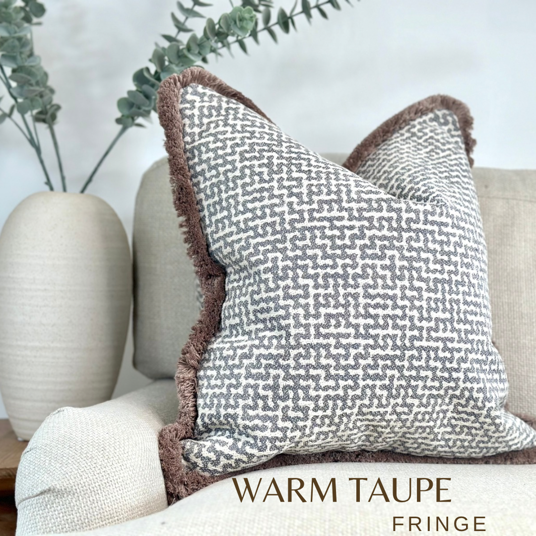 A luxury Grey Blue silver geometric cushion with beige fringe trim. Sitting on a neutral sofa with minimalist decor