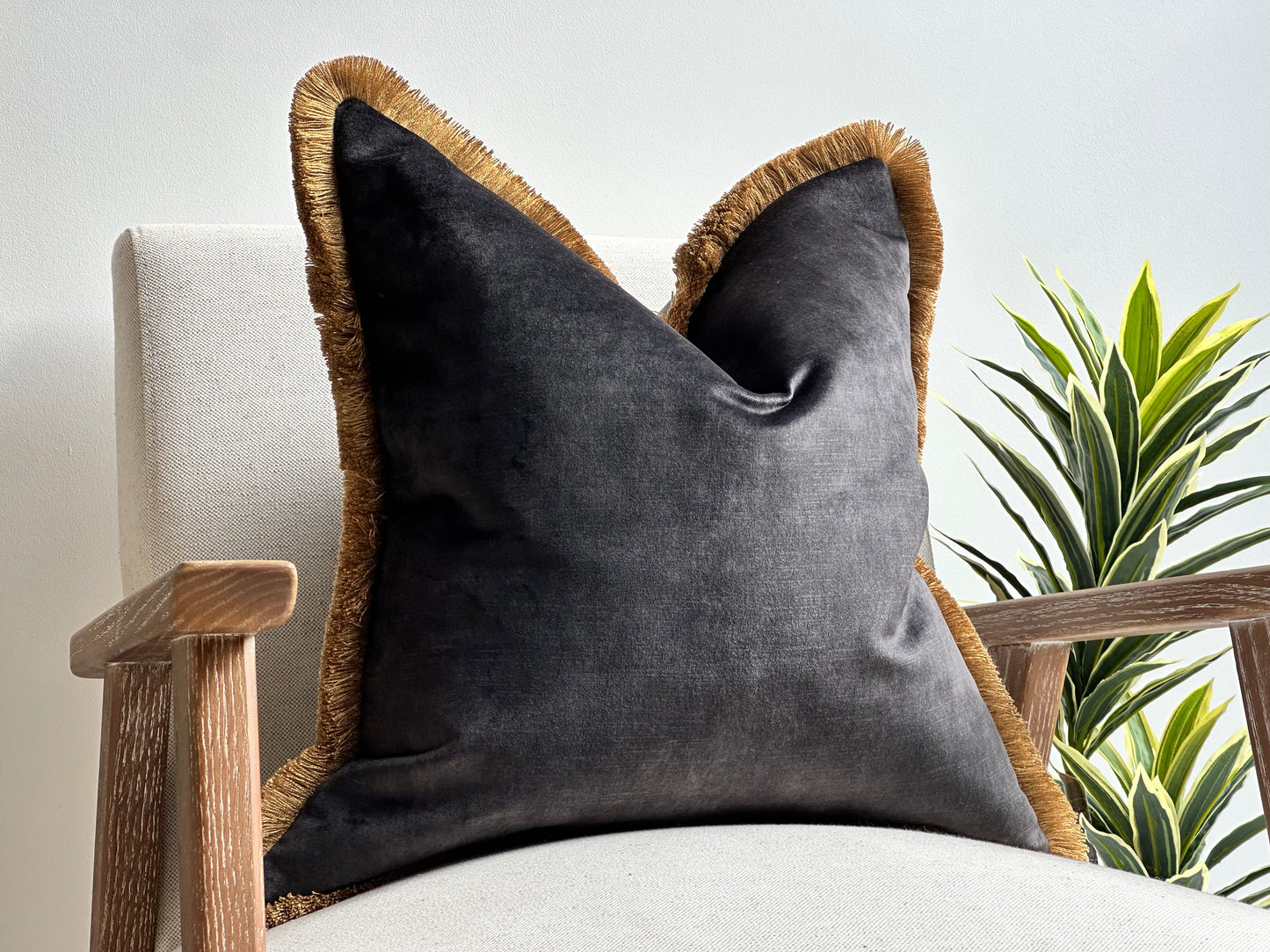 A luxury charcoal black velvet cushion with gold fringe trim placed on a neutral-toned armchair.