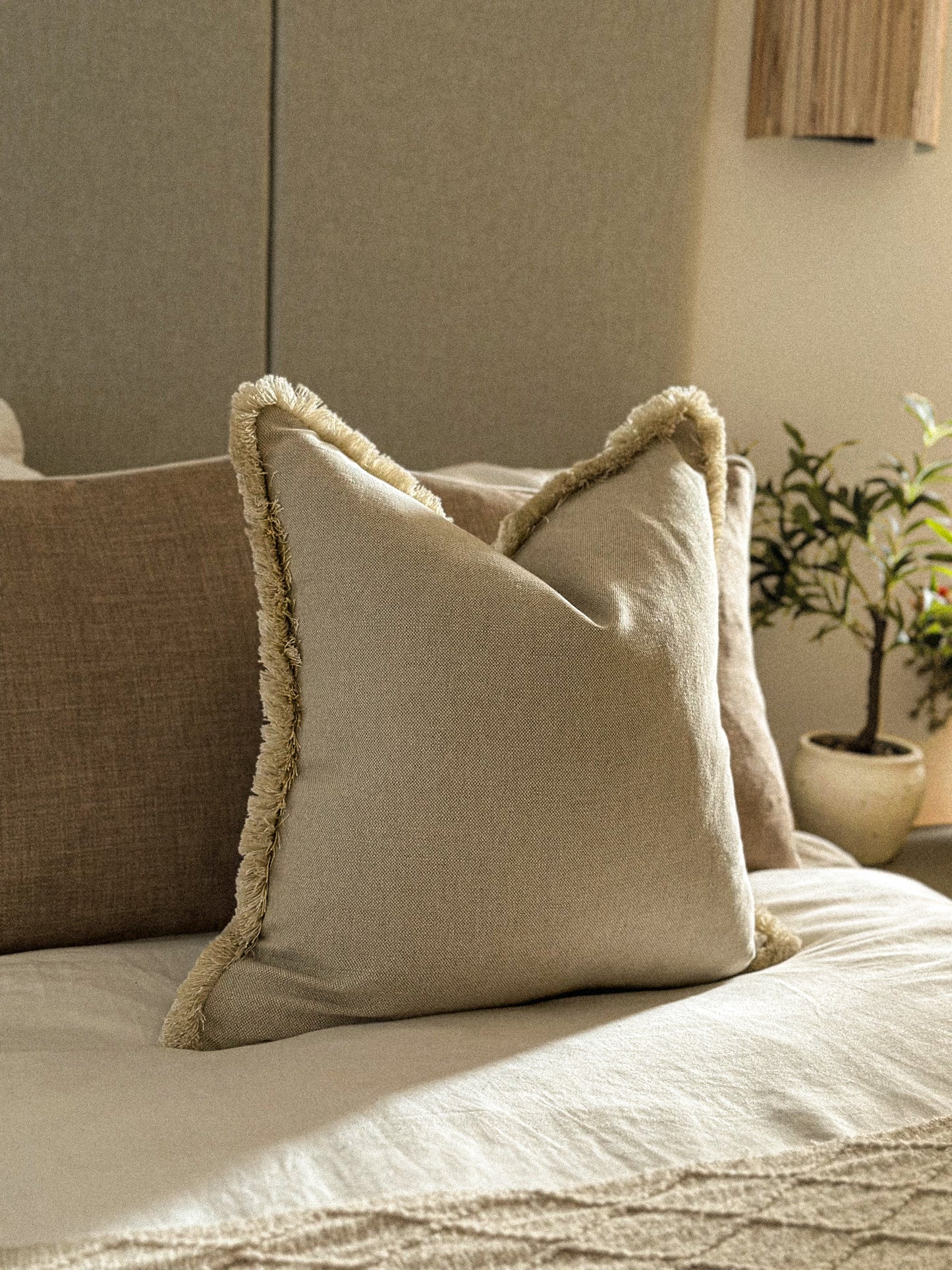 Earthy neutral cushion with fringe trim styled on a bed with warm tones.