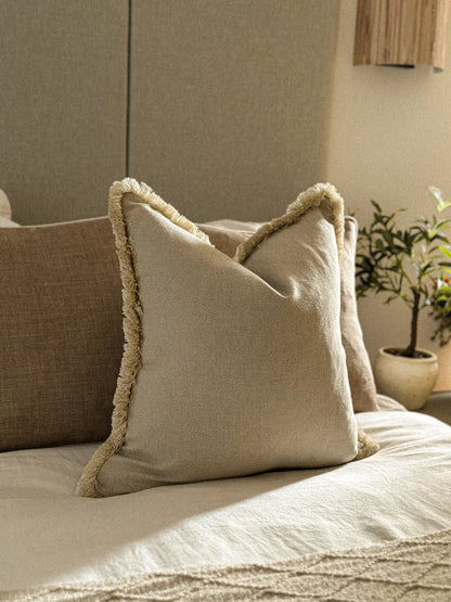 Earthy neutral cushion with fringe trim styled on a bed with warm tones.