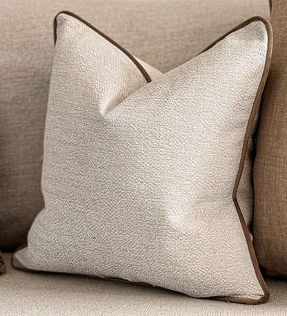 A square throw pillow in a textured cream fabric with beigepiping, resting on a beige upholstered sofa.