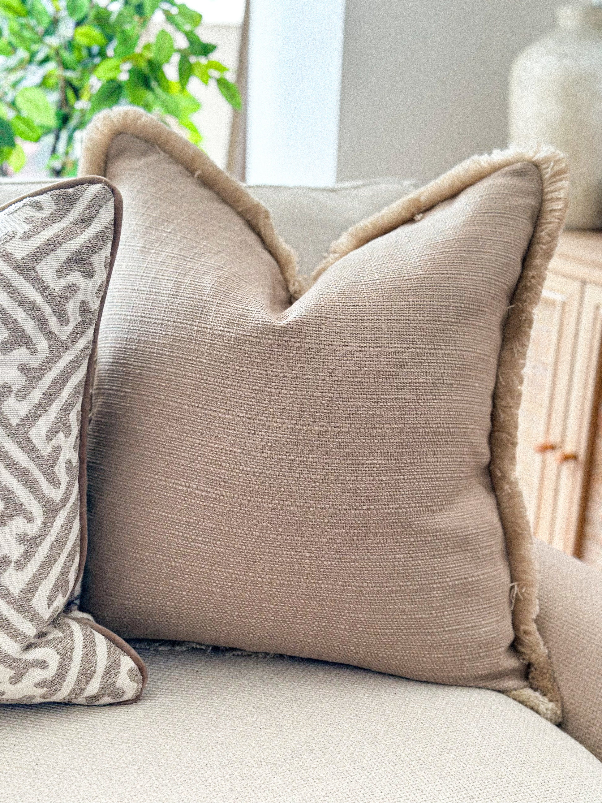 Neutral taupe textured cushion with fringe trim, styled on a cream sofa.