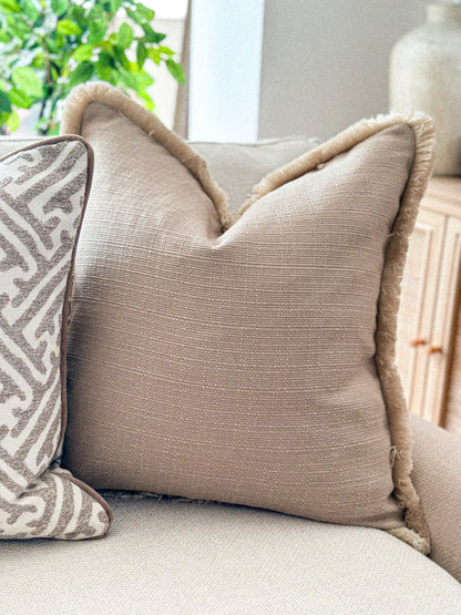 Neutral taupe textured cushion with fringe trim, styled on a cream sofa.