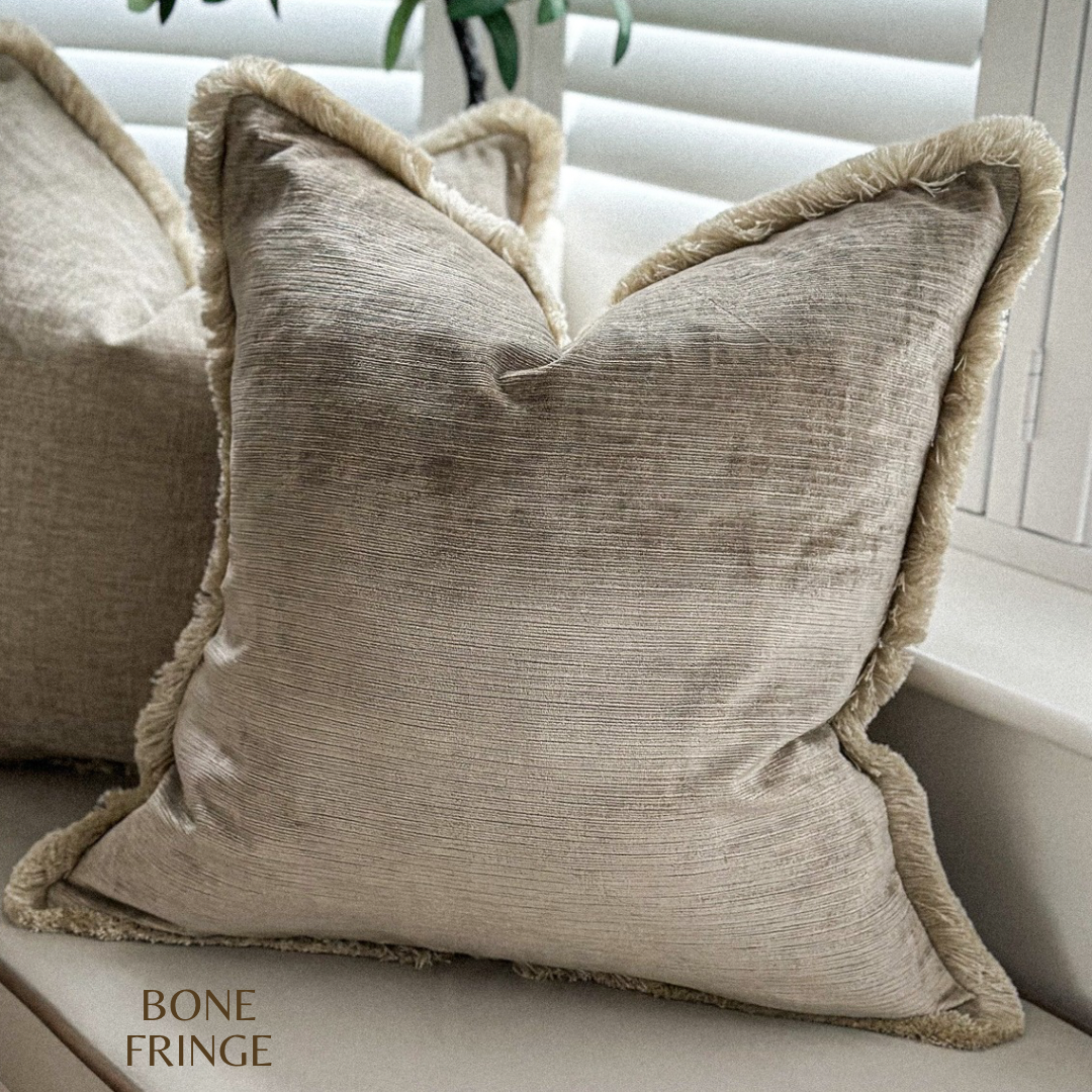 Soft beige velvet pillow cover with tonal fringe trim in a cosy living space.