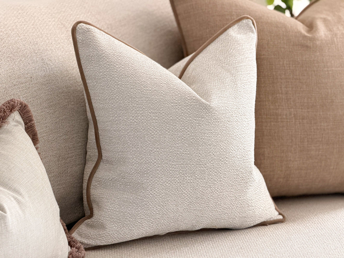 A square cushion in textured off-white fabric with a contrasting brown piped edge, styled on a beige sofa alongside other neutral-toned cushions.