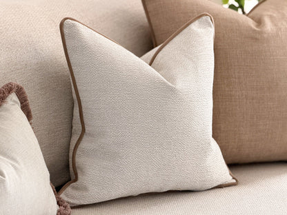 A square cushion in textured off-white fabric with a contrasting brown piped edge, styled on a beige sofa alongside other neutral-toned cushions.