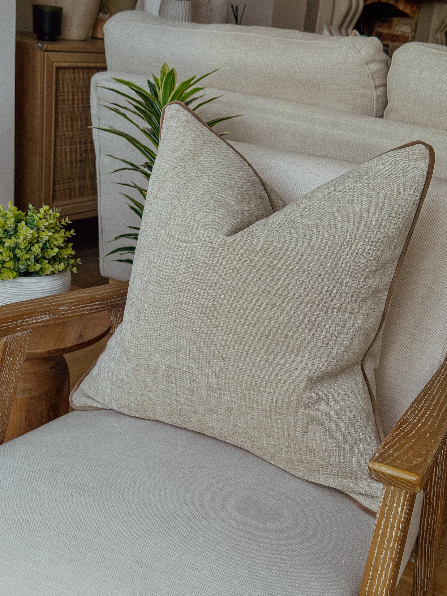 Beige linen-look cushion with taupe piping styled on a neutral armchair.”
