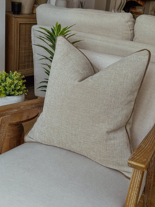 Beige linen-look cushion with taupe piping styled on a neutral armchair.”
