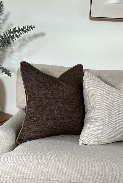 dark brown cushion cover with subtle textured fabric and mocha piping, styled on a neutral armchair beside a light herringbone pillow. Handmade, luxury feel.