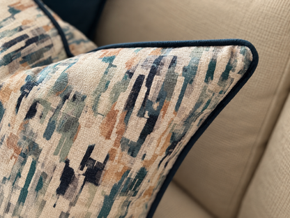 A close-up of a decorative cushion with an abstract geometric pattern in beige, teal, navy, and brown, finished with dark navy piped edges, resting on a beige textured sofa.
