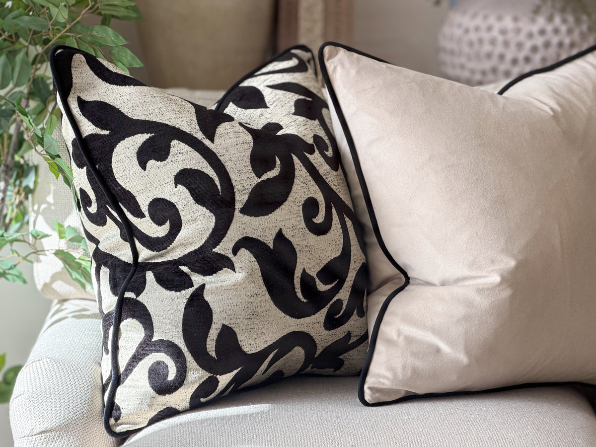 Two decorative cushions on a light-colored sofa. The front pillow has a black botanical pattern on a textured cream fabric, and the rear pillow is a solid cream velvet with black piping.