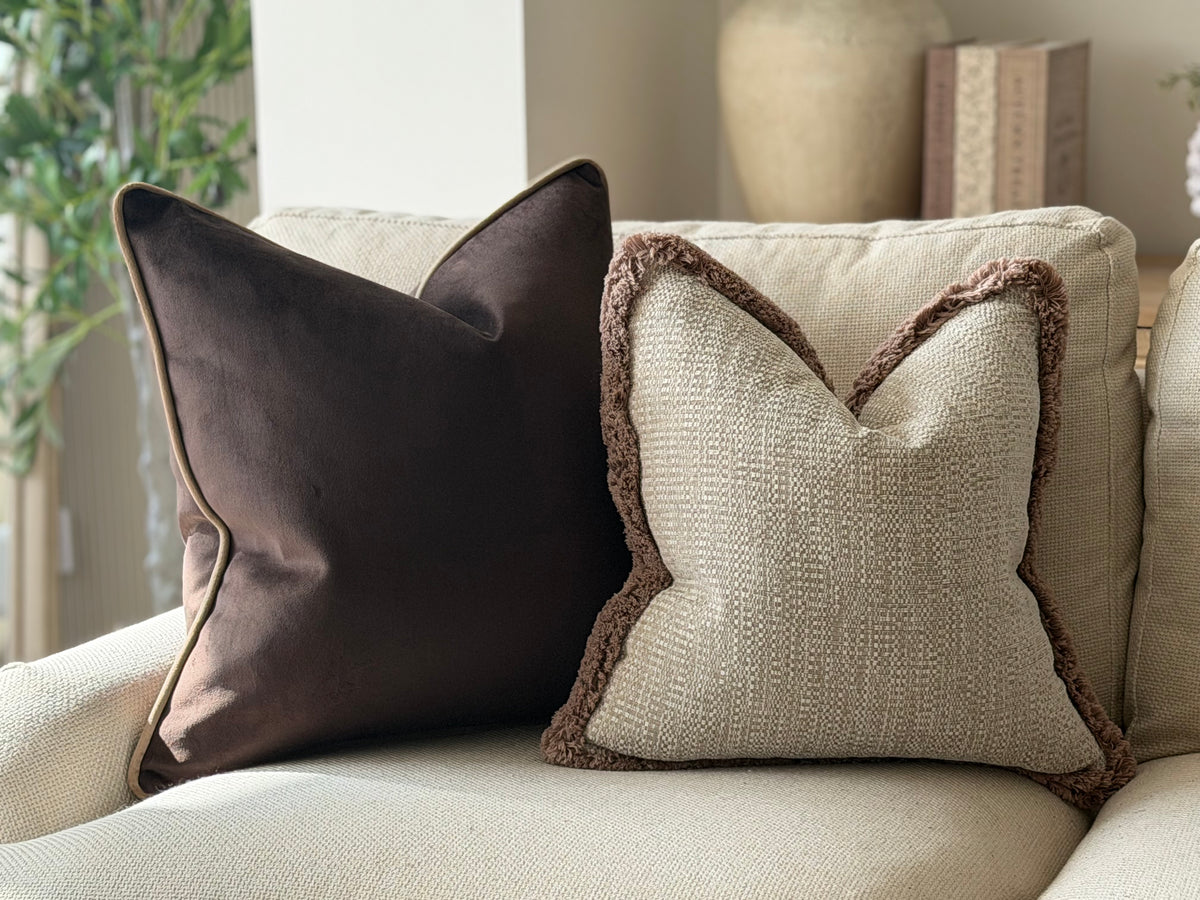 Two decorative cushions on a light beige sofa in a softly lit living room. One pillow is dark brown velvet with piped edges, and the other is a textured beige pillow with a brown fringe trim.