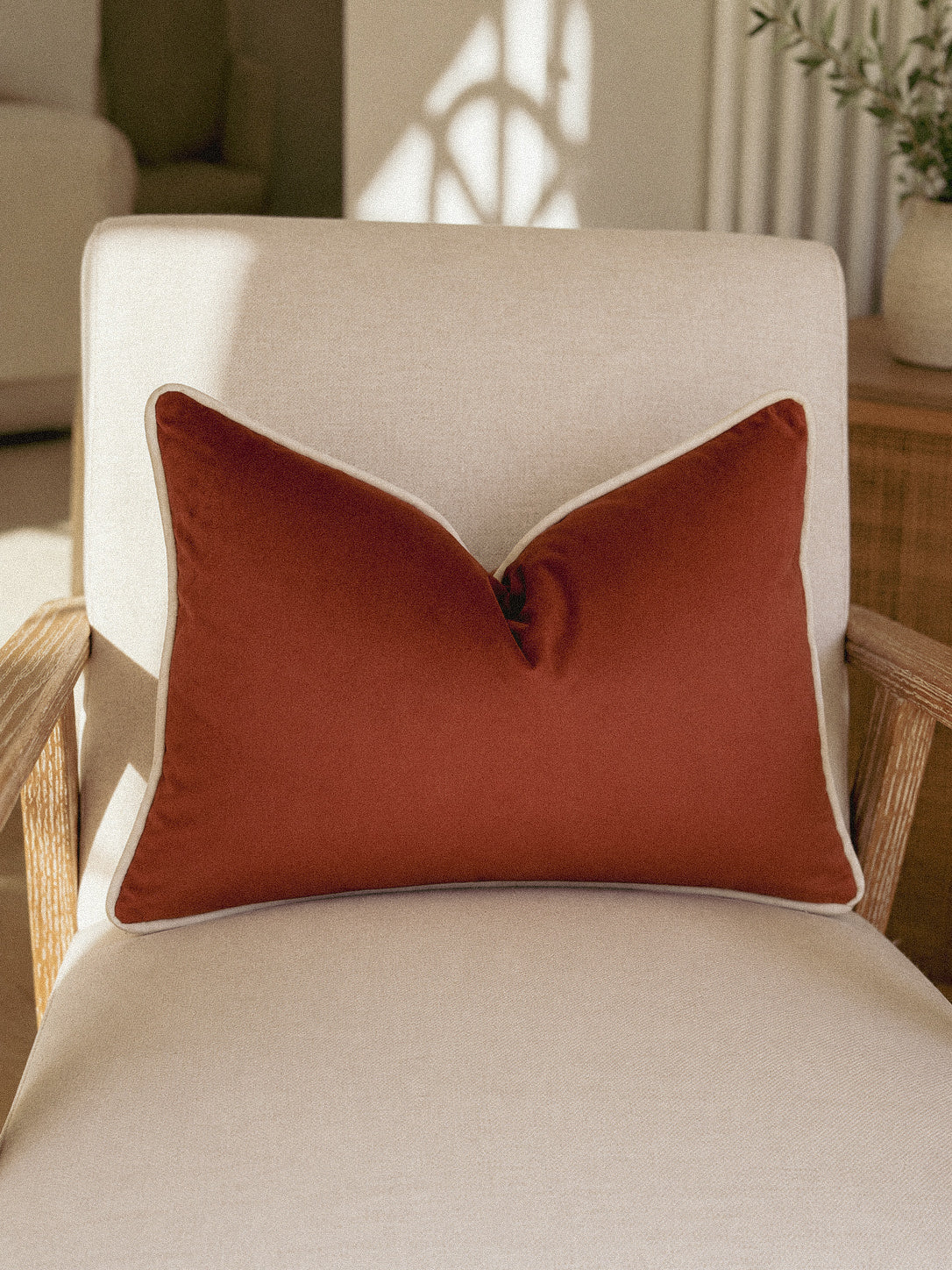 A rust-orange rectangular throw pillow with white piping, resting on a cream-colored armchair in a softly lit room.