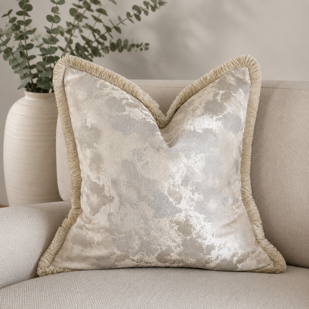 Champagne pearl decorative cushion with ivory fringe trim styled on a neutral textured armchair, featuring a soft metallic designer fabric in elegant champagne and ivory tones.