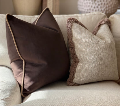 Two decorative throw pillows on a light beige sofa. One pillow is dark brown with light beige piping, and the other is beige with a brown textured trim.