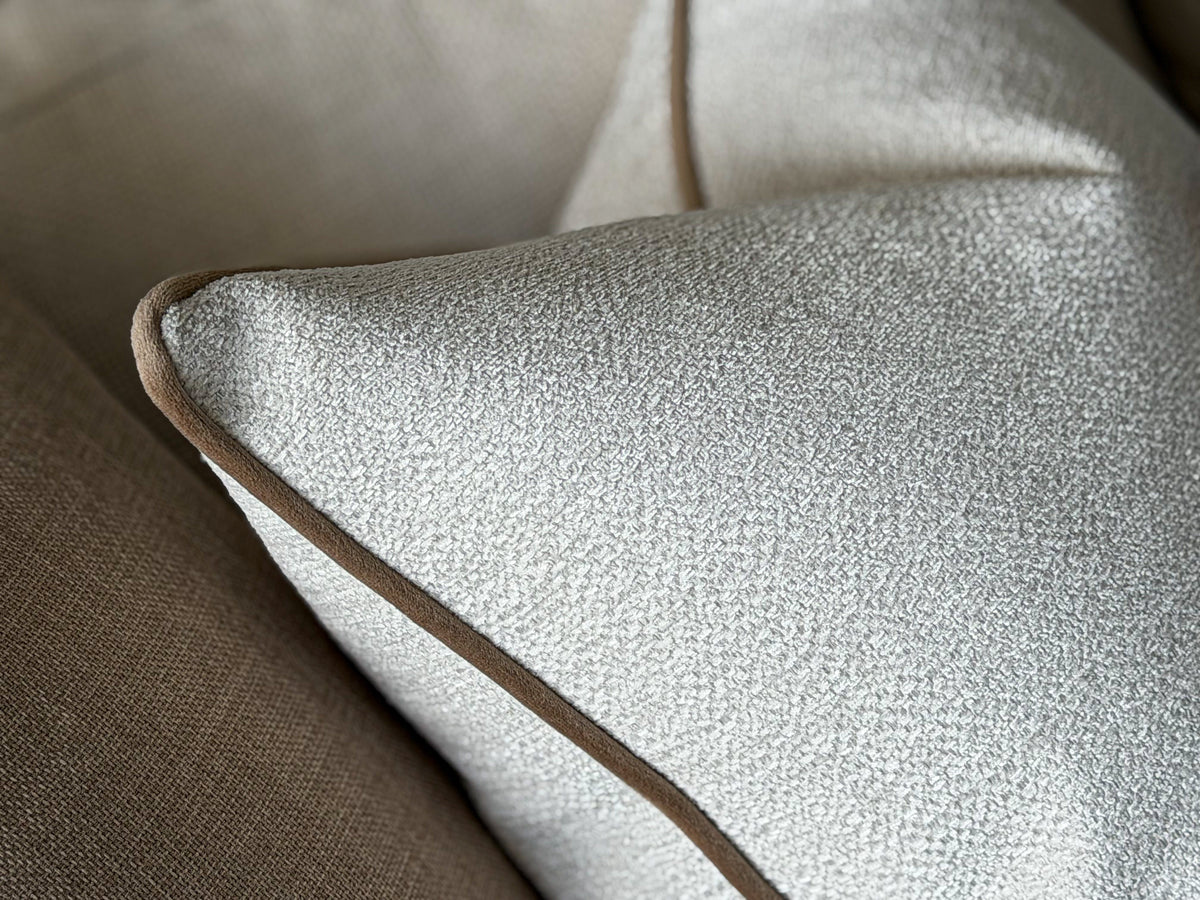 Close-up of a textured ivory throw pillow with a contrasting brown piping edge, resting on a neutral beige sofa.