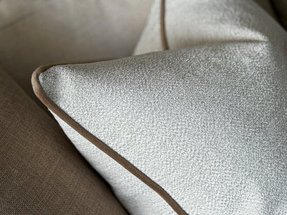Close-up of a textured ivory throw pillow with a contrasting brown piping edge, resting on a neutral beige sofa.