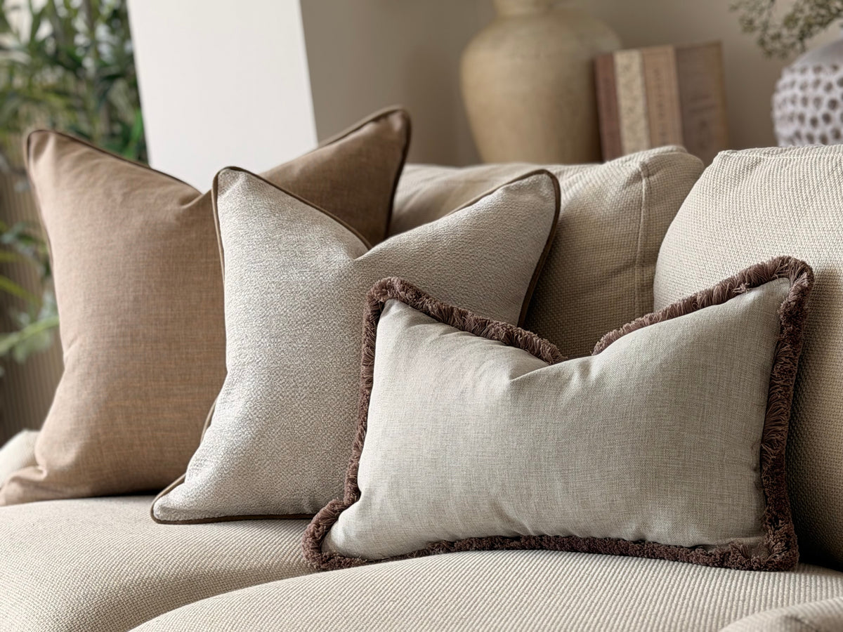 A set of neutral-toned throw pillows arranged on a beige sofa in a softly styled living room. The pillows include solid taupe, light beige, and a light gray pillow with a dark brown fringed edge.