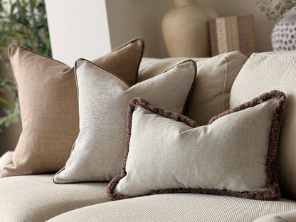 A set of neutral-toned throw pillows arranged on a beige sofa in a softly styled living room. The pillows include solid taupe, light beige, and a light gray pillow with a dark brown fringed edge.