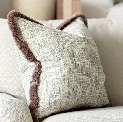 A square throw pillow with a textured cream fabric and a contrasting beige taupe plush trim, resting on a light beige sofa.