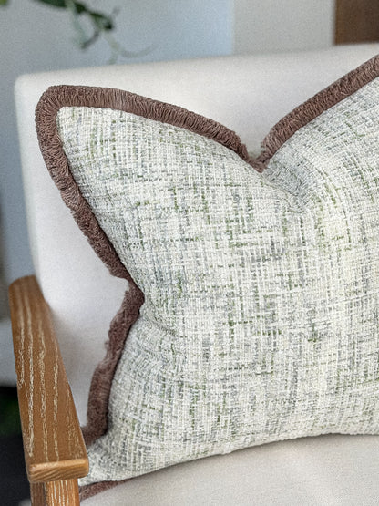 A close-up of a textured throw pillow with a woven light beige and green pattern and a contrasting brown taupe fringed edge,