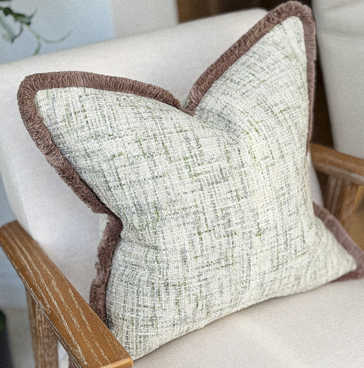 A square decorative cushionwith a textured cream and light green woven fabric body and a contrasting brown fringed edge, resting on a light-colored upholstered armchair with wooden arms.