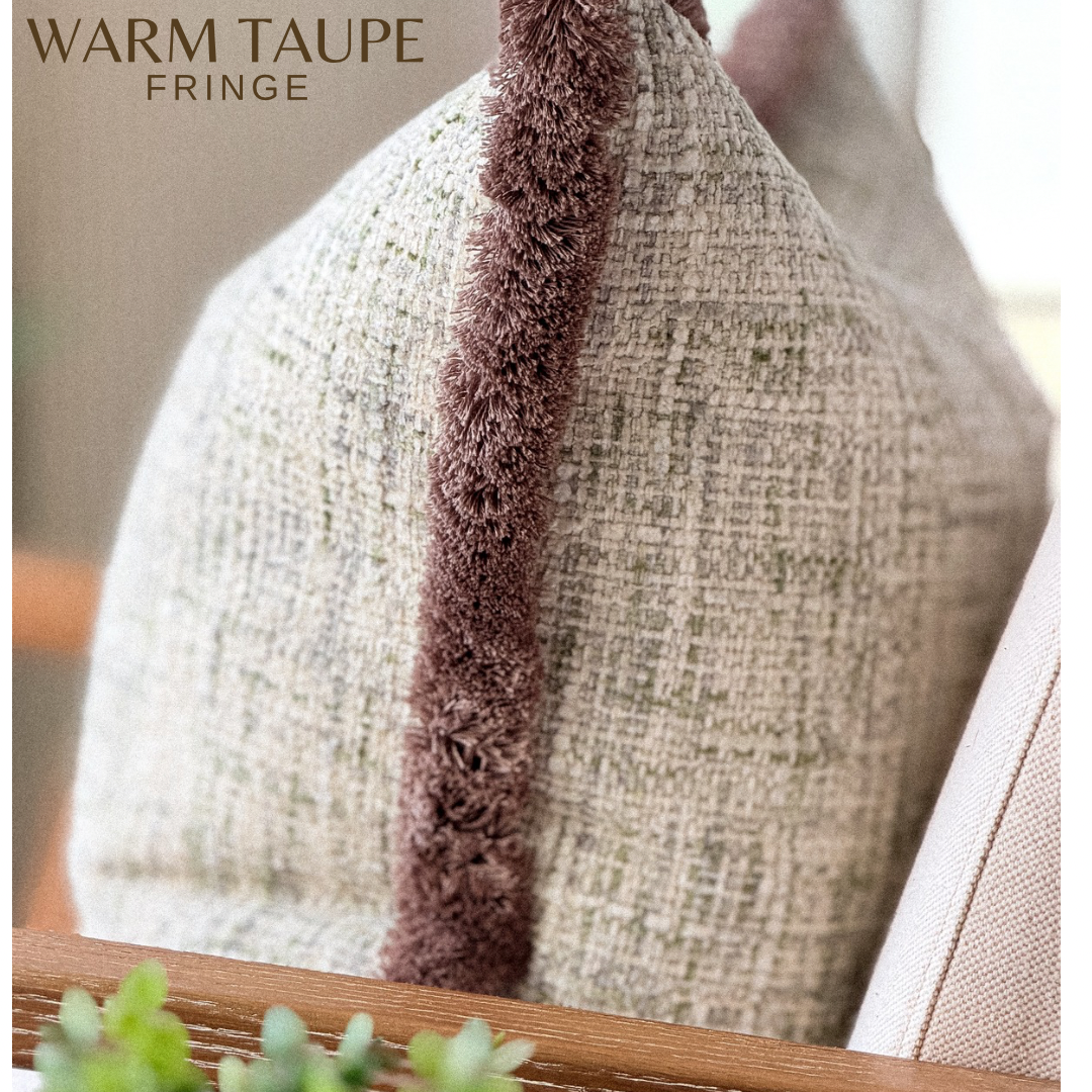 A close-up of a textured throw pillow in warm taupe woven fabric with a prominent vertical brown faux-fur fringe trim. The words 'WARM TAUPE FRINGE' are printed in the top left corner.