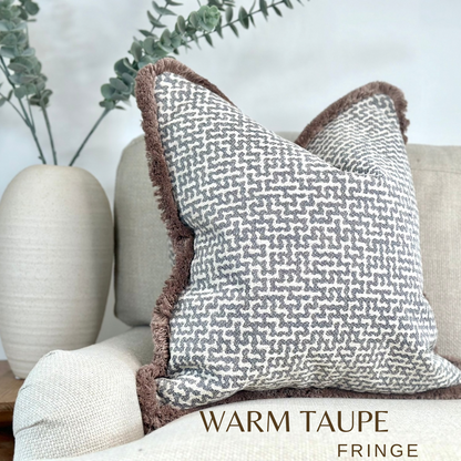 A luxury Grey Blue silver geometric cushion with beige fringe trim. Sitting on a neutral sofa with minimalist decor