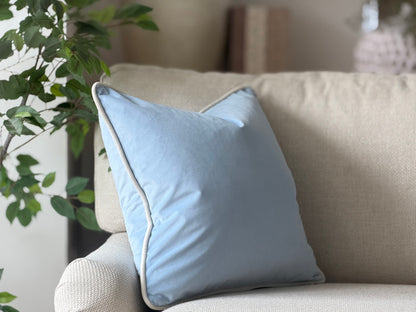 A light blue velvet square cushion with white piping trim.
