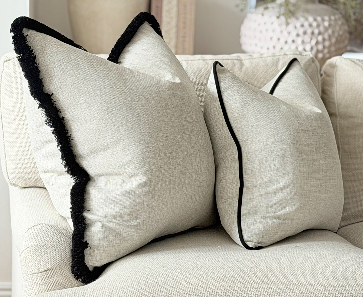A photo of 2 cushions in a natural linen like fabric one has a black fringe trim and the other is finished with a black velvet piping.