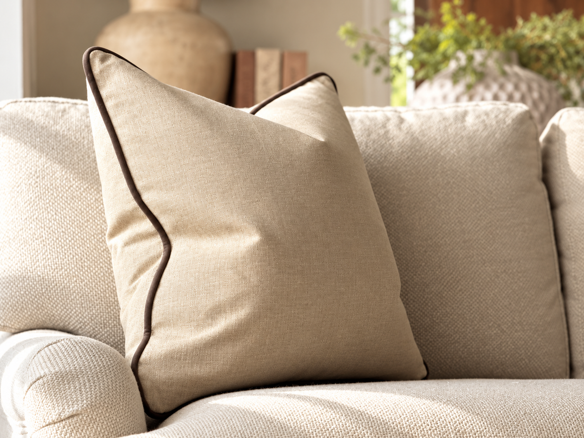 A square beige throw pillow with dark brown piping trim, resting on a textured light beige sofa in a softly lit living room setting.