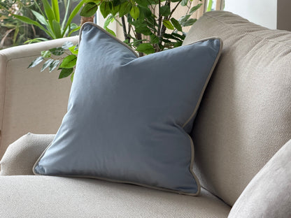 A pastel light blue square throw pillow with a ivory piped edge, resting on a beige textured sofa with green plants in the background.