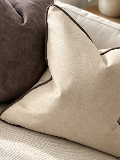 A close-up of a stone beige textured throw pillow with dark brown piping, resting on a light beige sofa next to a dark brown pillow.