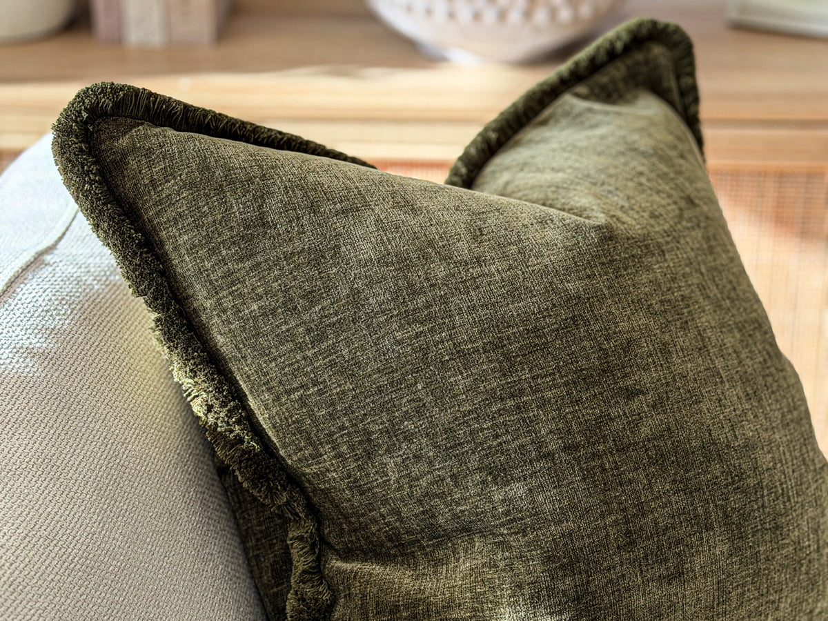 A close-up of a textured olive green throw pillow with a fringed edge, resting on a light-colored sofa.