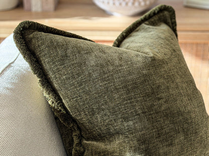 A close-up of a textured olive green throw pillow with a fringed edge, resting on a light-colored sofa.