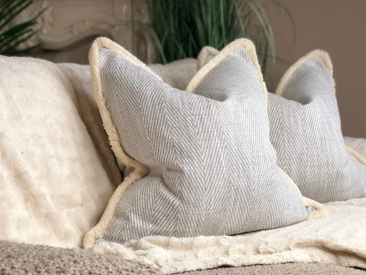 Light herringbone cushion cover with soft cream fringe trim, styled on a beige faux fur throw. Square neutral pillow with subtle chevron pattern and warm, inviting texture.